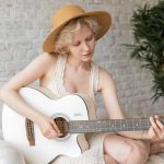 Focused female in hat and dress playing acoustic guitar while sitting on armchair near potted plant during rehearsal at home